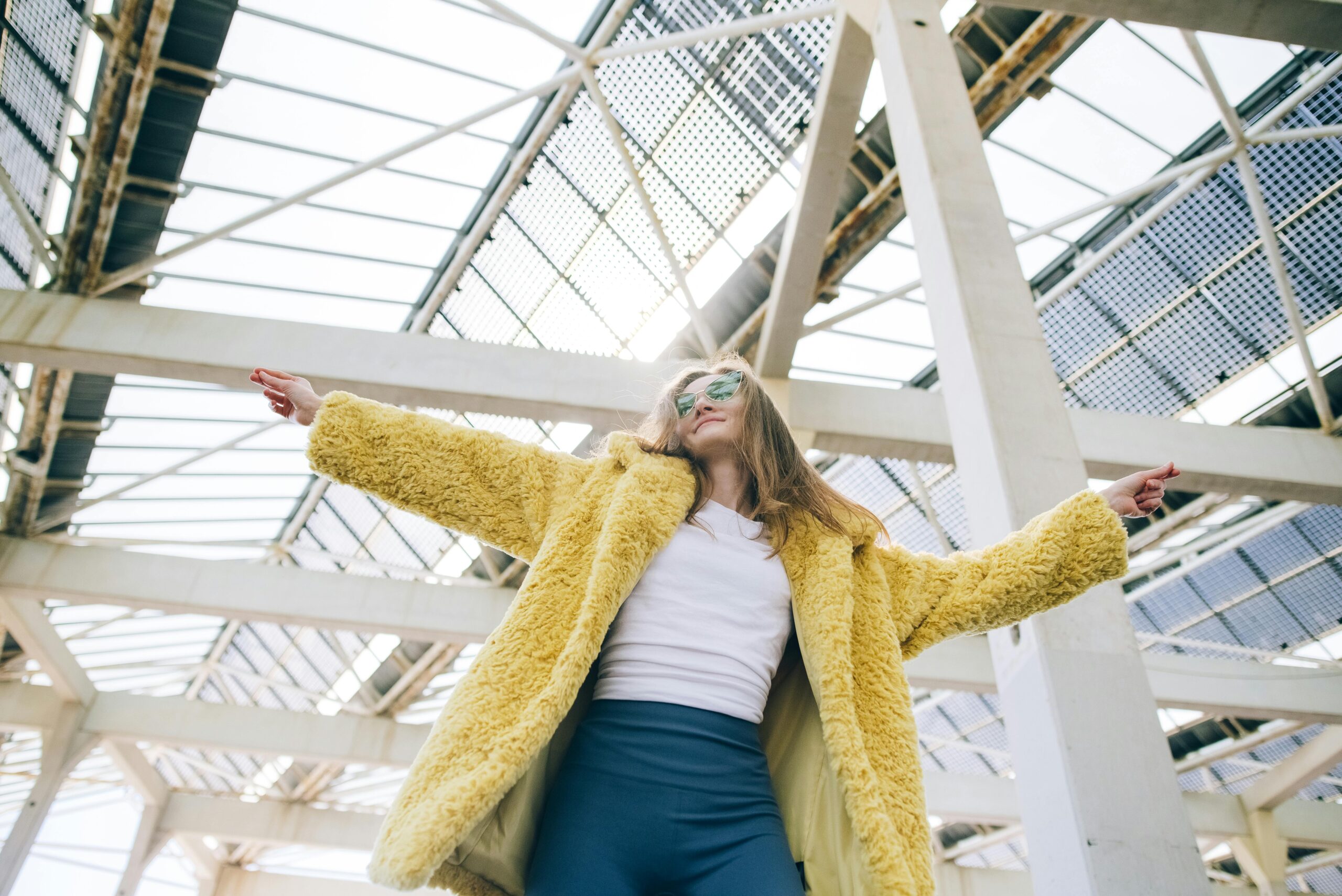 Fashionable woman in a yellow faux fur coat posing confidently under a modern structure.